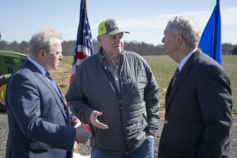 Three men with flag and farm field in background.