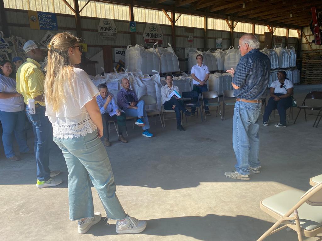 People in a delegation from Argentina, Paraguay, and Uruguay stand in a barn and sit front of bags of cover crop seed while listening to Bobby Hutchison discuss conservation efforts on his 130-year-old family farm.