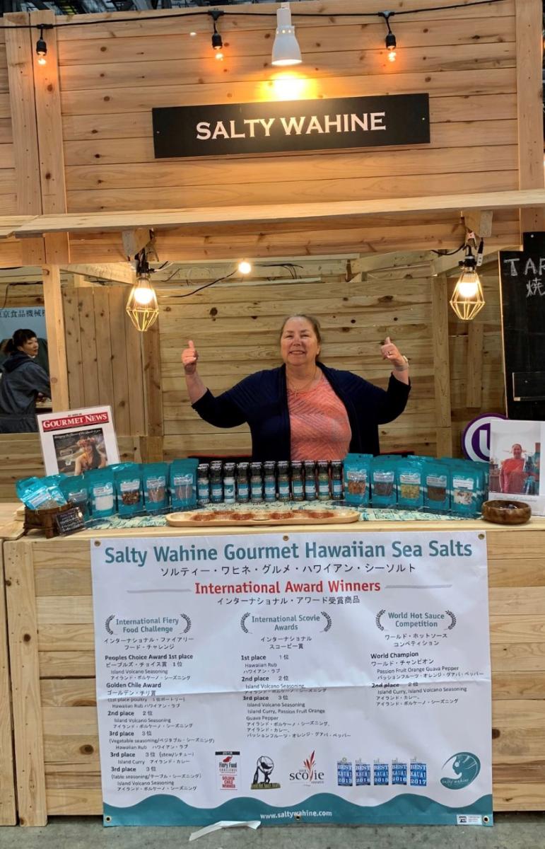 Woman-business owner, Laura Cristobal-Andersland, is standing behind her wooden booth and pavilion, displaying her gourmet seasonings and spices at a trade show.