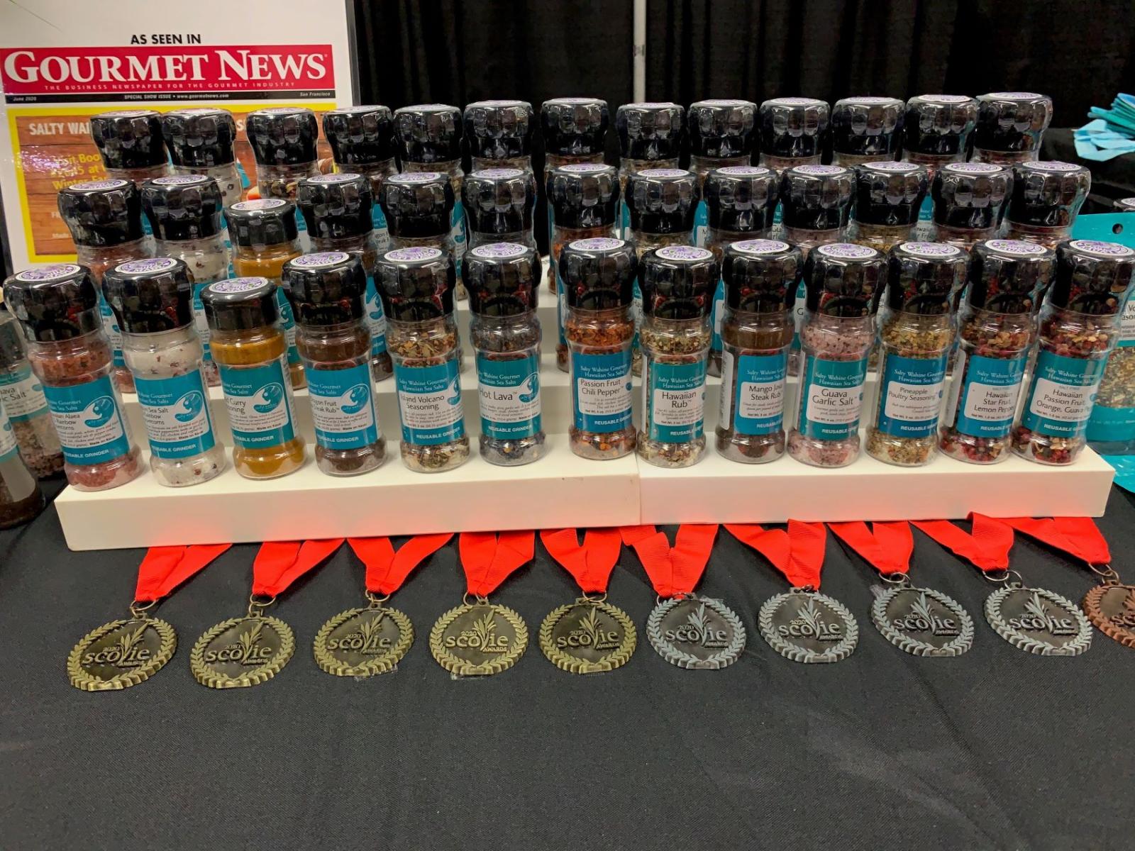 An array of gourmet spices made by Salty Wahine Gourmet Sea Salts are arranged on a three-level stand, with medals placed in front of the stand.