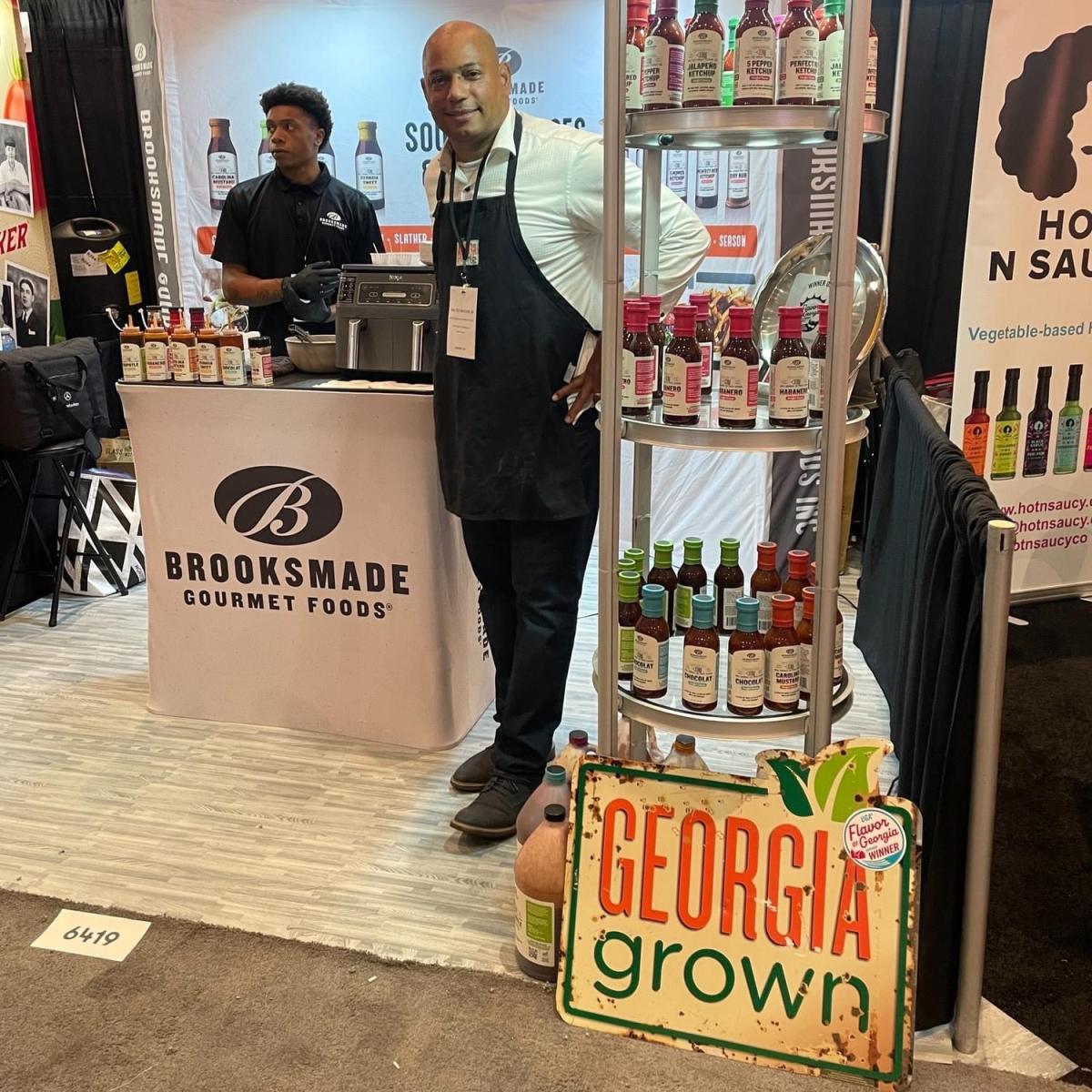 A gentleman in an apron poses in front of a booth with a gentleman standing behind the booth.