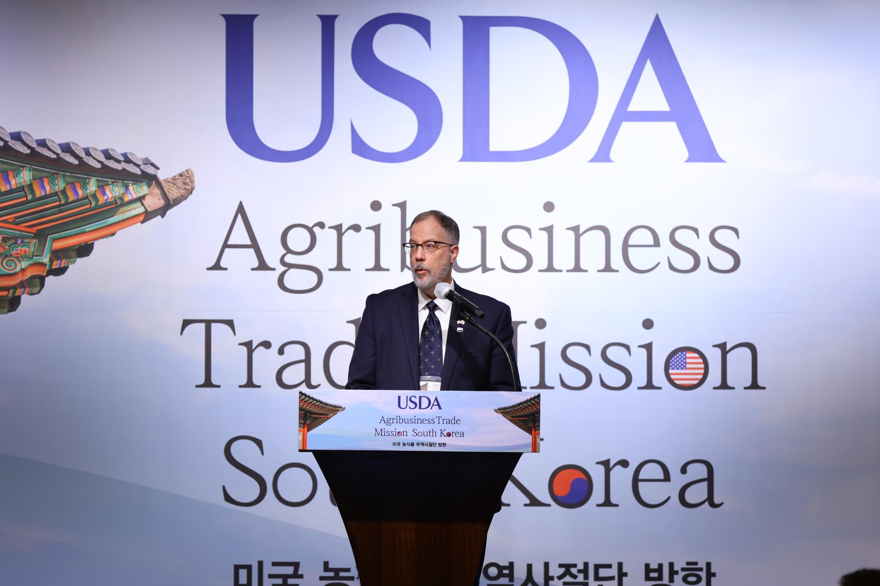 A man speaks at a podium with a background that reads USDA Agribusiness Trade Mission South Korea