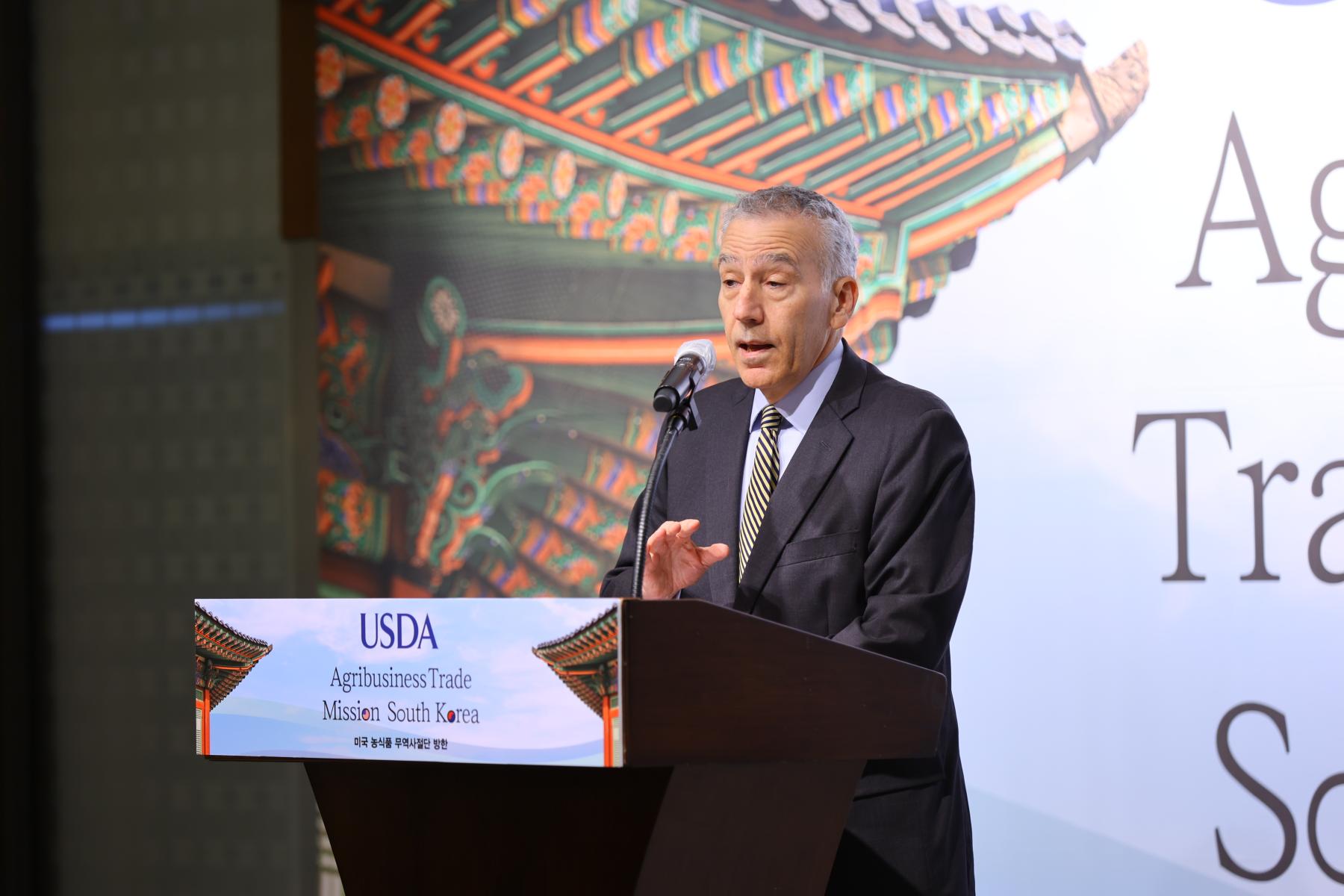 A man speaks at a podium with a label that reads USDA Agribusiness Trade Mission South Korea